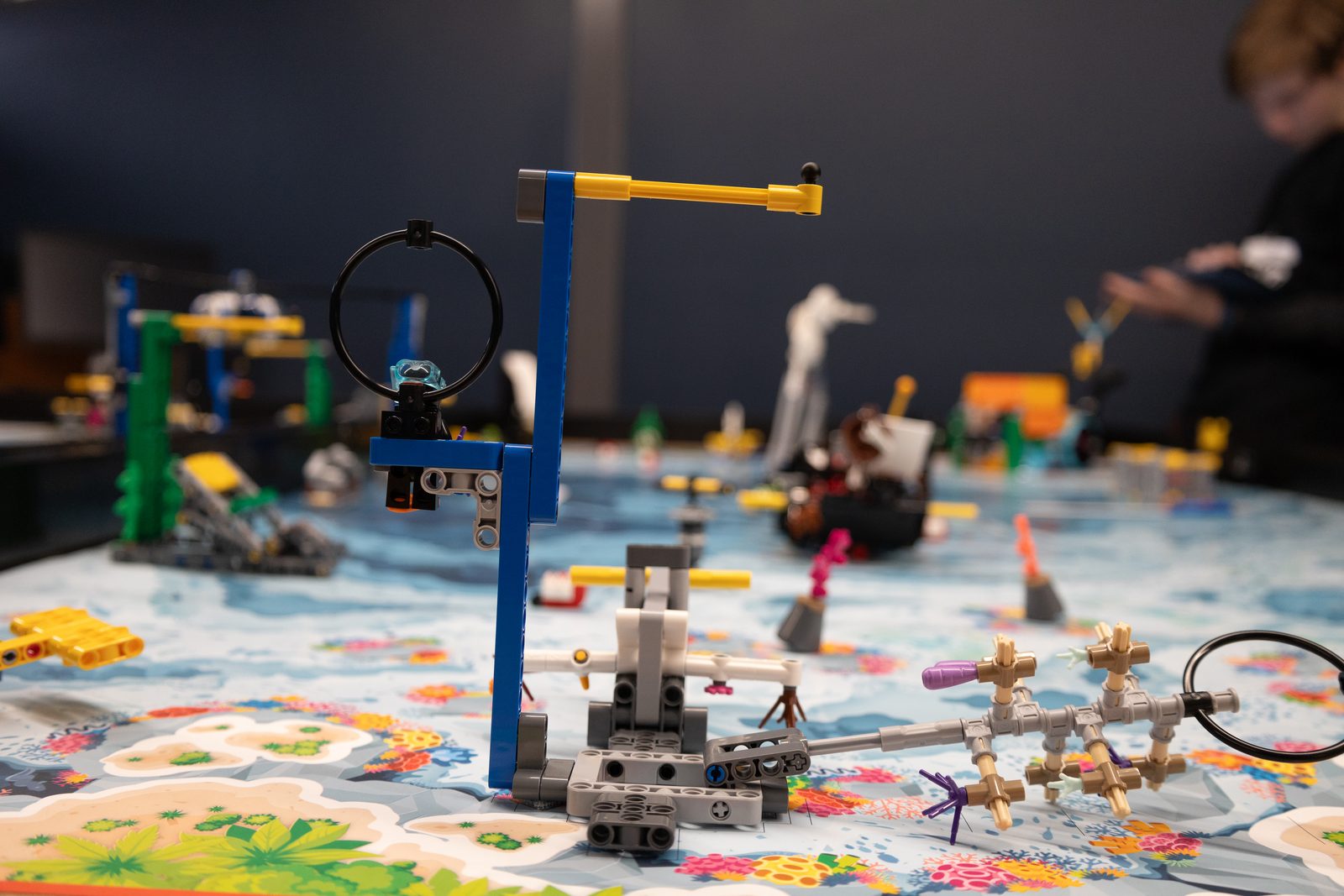 A student-built LEGO robot attachment on a FIRST LEGO League game table, with other teams' robots in the background